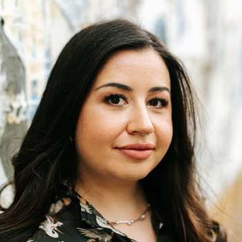 Woman with long dark hair standing beside textured wall, looking at camera.